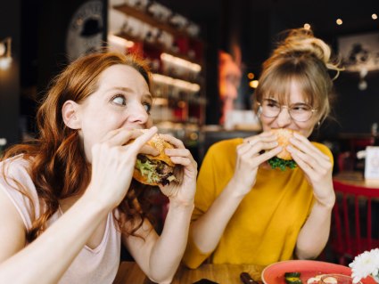Two female friends eating burger in a restaurant, © Westend61